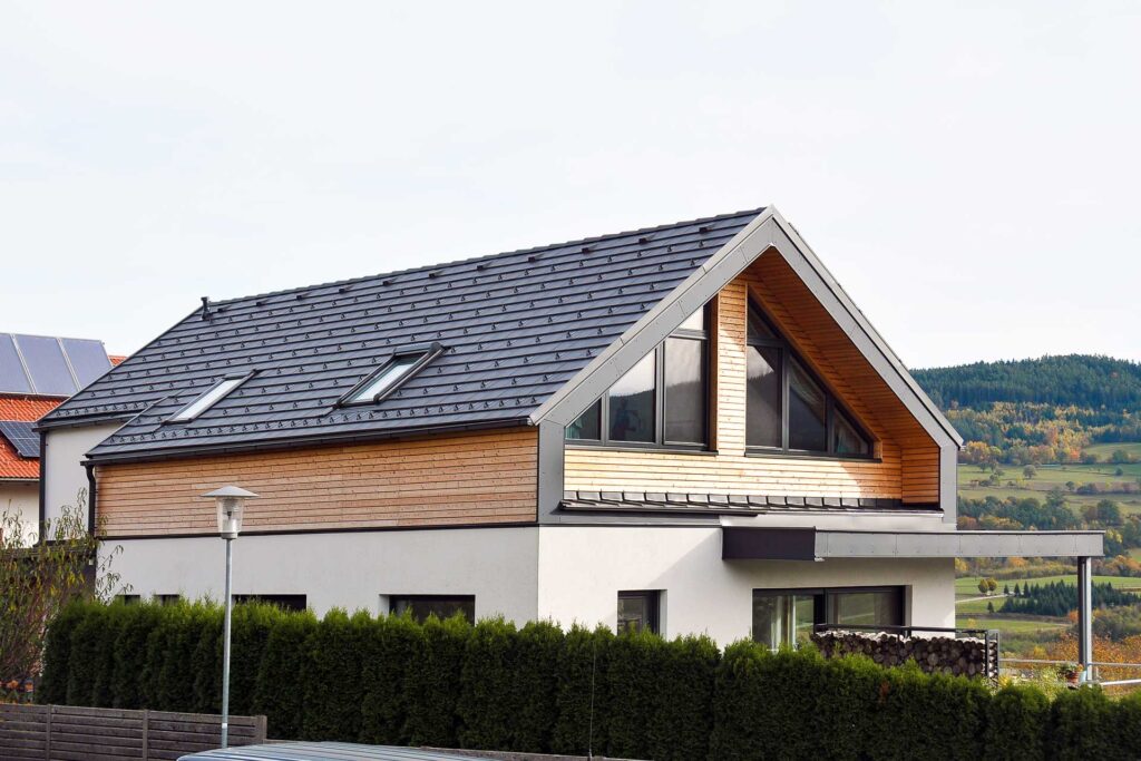 Modernes zweistöckiges Haus mit dunklem Schrägdach, großen Fenstern und Holzverkleidung im Obergeschoss. Das Haus ist von einer hohen grünen Hecke umgeben und blickt auf eine malerische, hügelige Landschaft im Hintergrund.