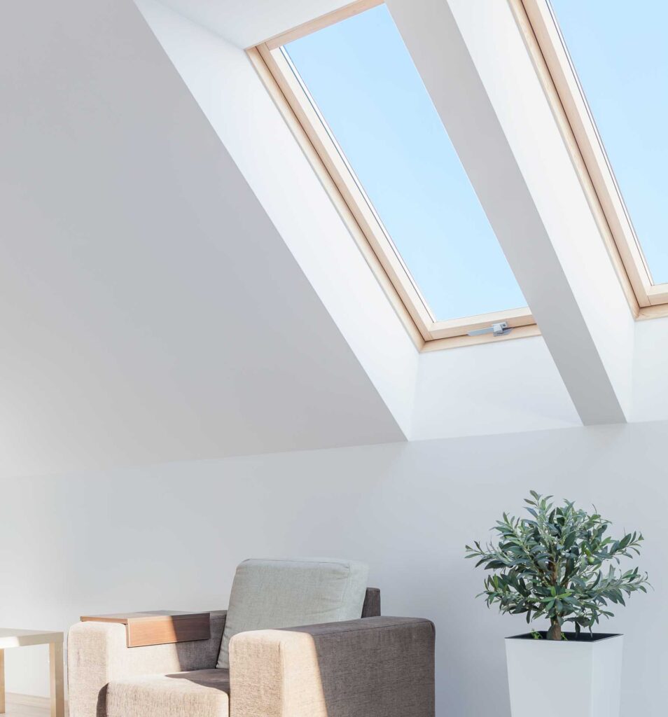 Ein modernes, minimalistisches Zimmer mit einem beigen Sessel, einem kleinen Beistelltisch, einer Topfpflanze und zwei großen Dachfenstern, die viel Tageslicht hereinlassen.