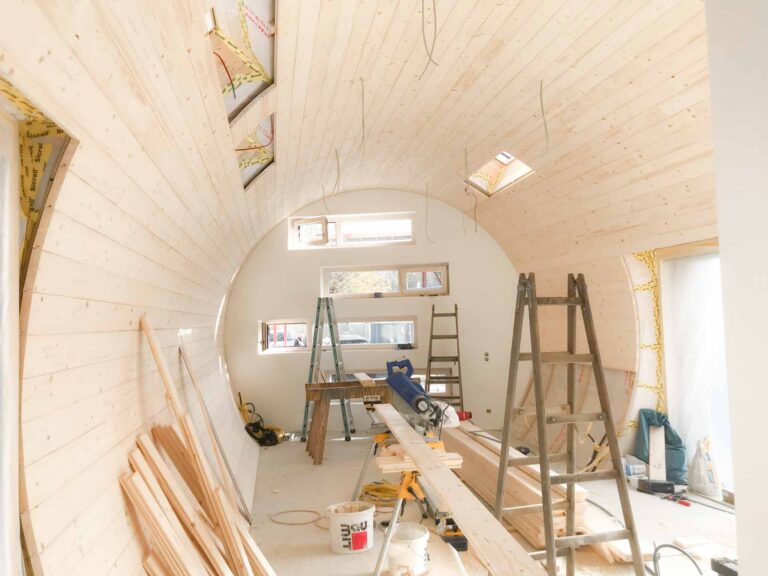 Ein teilweise gebauter Raum mit gebogenen Holzwänden, mehreren Leitern, Bauwerkzeugen, Holzbrettern und sichtbarer Isolierung. Mehrere rechteckige Fenster lassen natürliches Licht in den Arbeitsbereich.