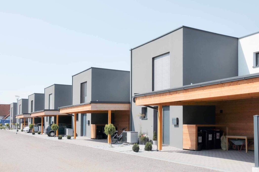Eine Reihe moderner Flachdachhäuser mit grauer Fassade und hölzernen Akzenten, jedes mit einem Carport, einige mit geparkten Autos, Pflanzen und Fahrrädern, unter einem klaren blauen Himmel.
