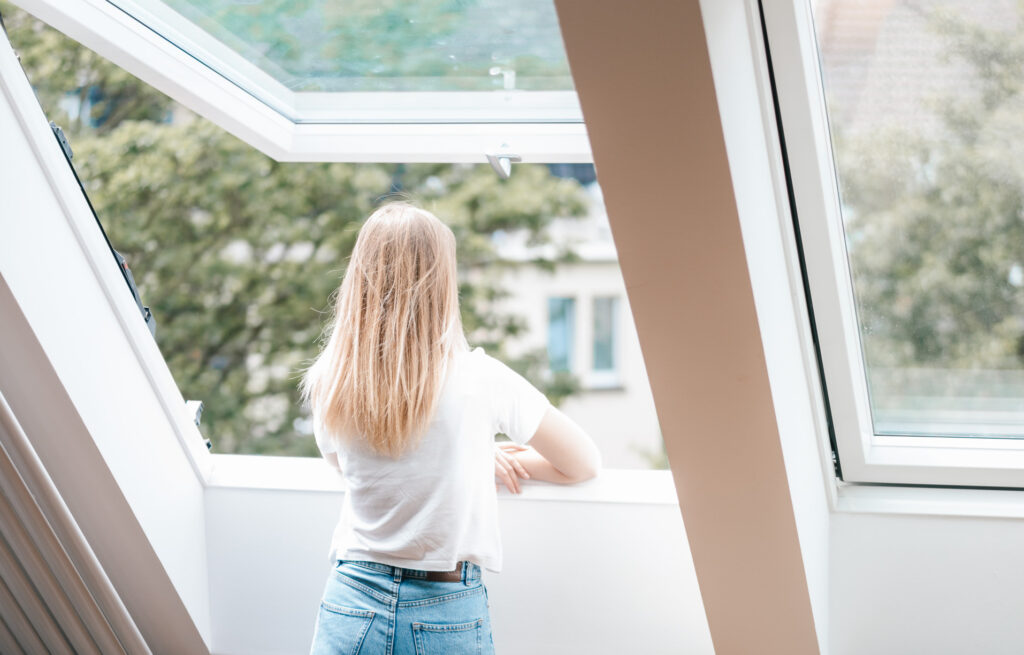 Eine Person mit langen blonden Haaren, die ein weißes Hemd und Jeans trägt, steht in einem hellen, modernen Raum am offenen Fenster und blickt auf Bäume und Gebäude hinaus.