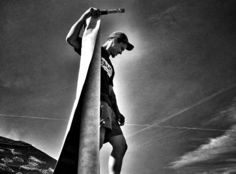 Eine Person mit Mütze und Shorts steht auf einem Dach und hält ein großes zusammengerolltes Tuch und ein Werkzeug vor einem dramatischen, bewölkten Himmel in Schwarz-Weiß.