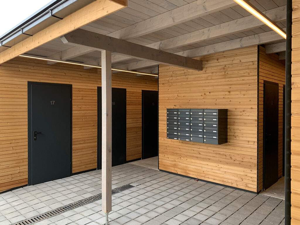 Ein moderner Innenhof mit hellen Holzwänden, grauen Türen mit den Nummern 17, 18 und 19, einer Reihe grauer Briefkästen an der Wand und einem gefliesten Boden unter einem überdachten Dach, das auch als Carport dient.